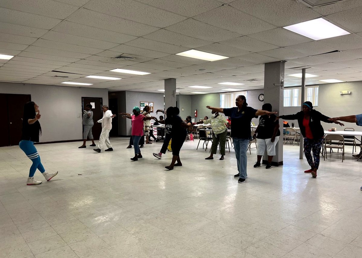 A Senior Friendly Fitness class in a Glades community hall, standing in a loose circle with arms extended as an instructor leads from the front